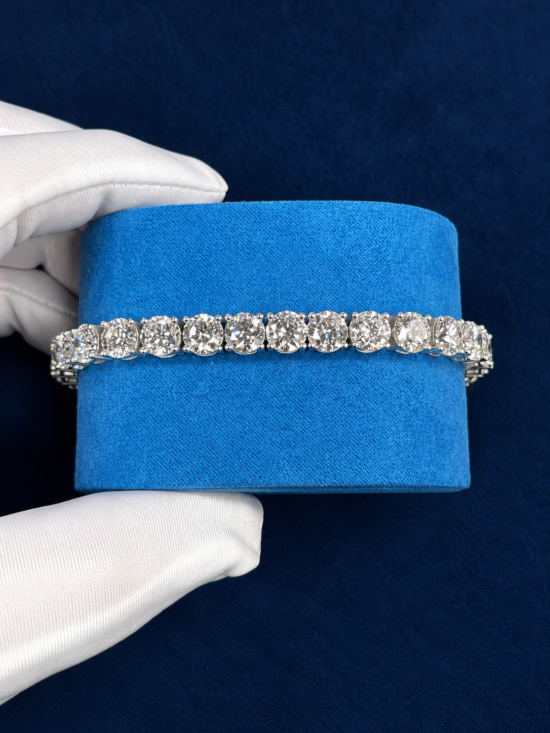 Custom iced tennis bracelet laid straight across a blue velvet cushion, round moissanite stones in four-prong settings, held in a white glove