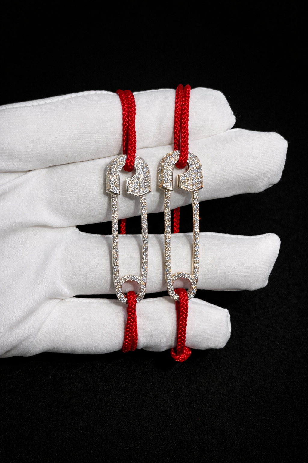 Custom icon bracelet with a fully iced safety-pin motif on a braided red cord, held in a white glove against a black background