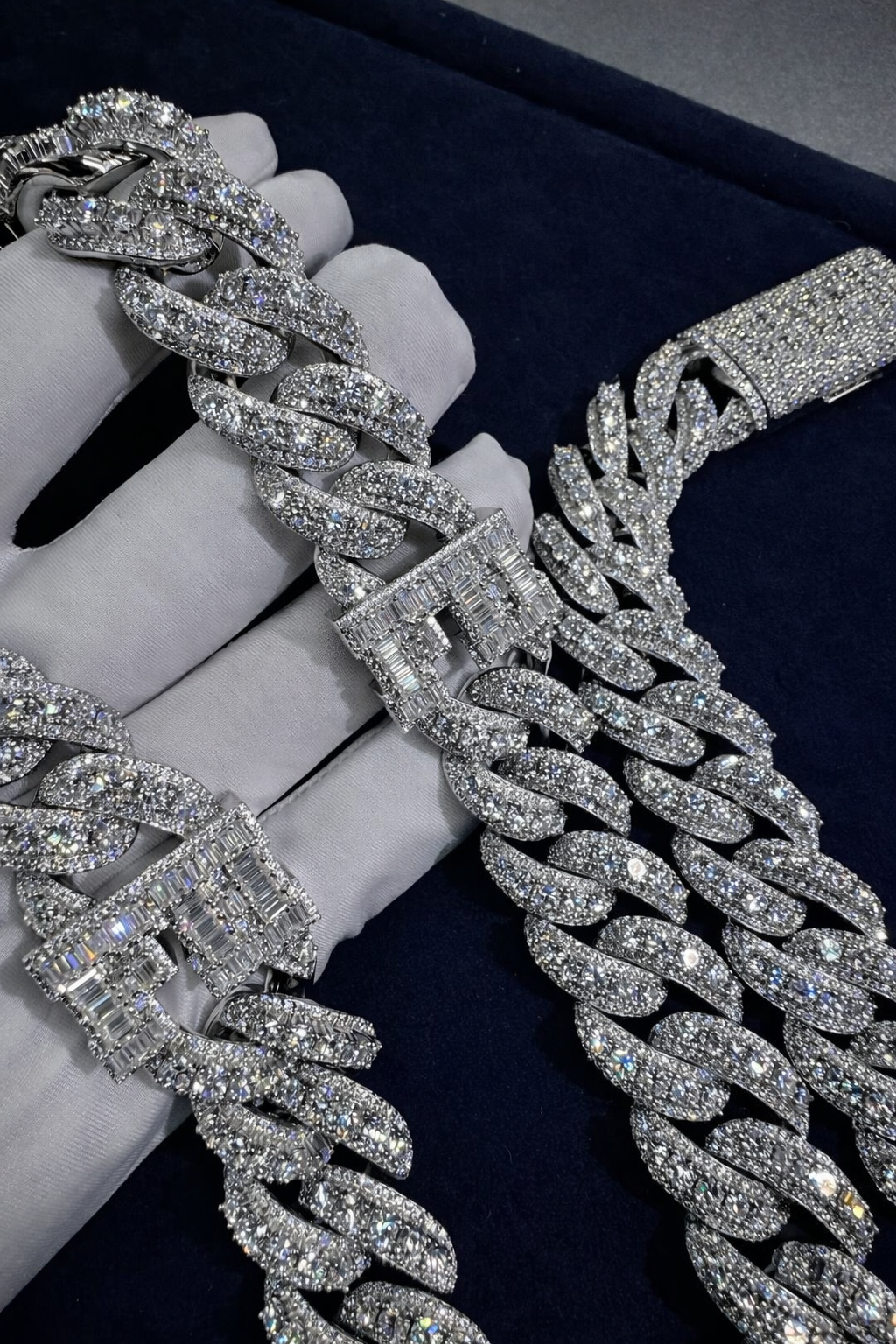 Macro close-up of two baguette-set letter motifs on the fully iced icon chain, held across a white-gloved hand