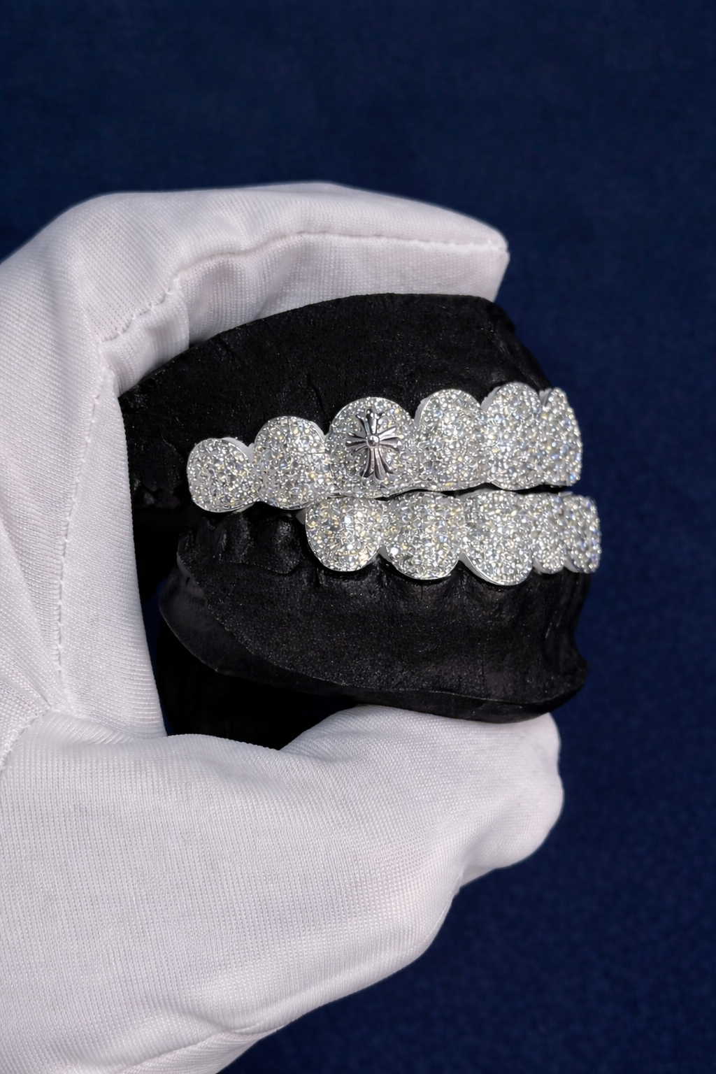 Custom iced grillz set with a raised cross emblem on the upper row, paved with moissanite, fitted on a black dental mould held in a white glove against a navy background