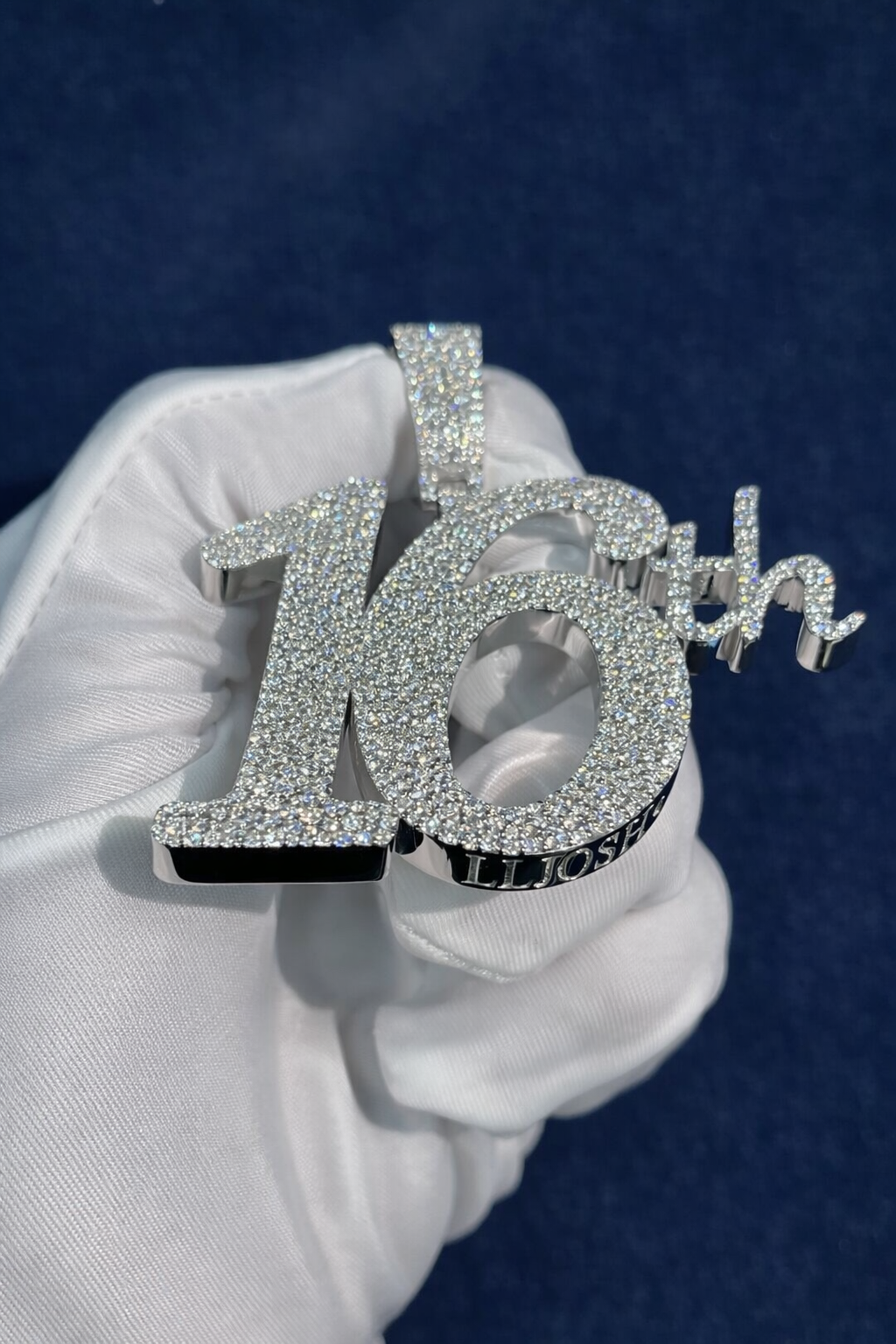 Custom iced 16th birthday pendant held upright in a white glove, bold serif numeral paved with moissanite against a navy background