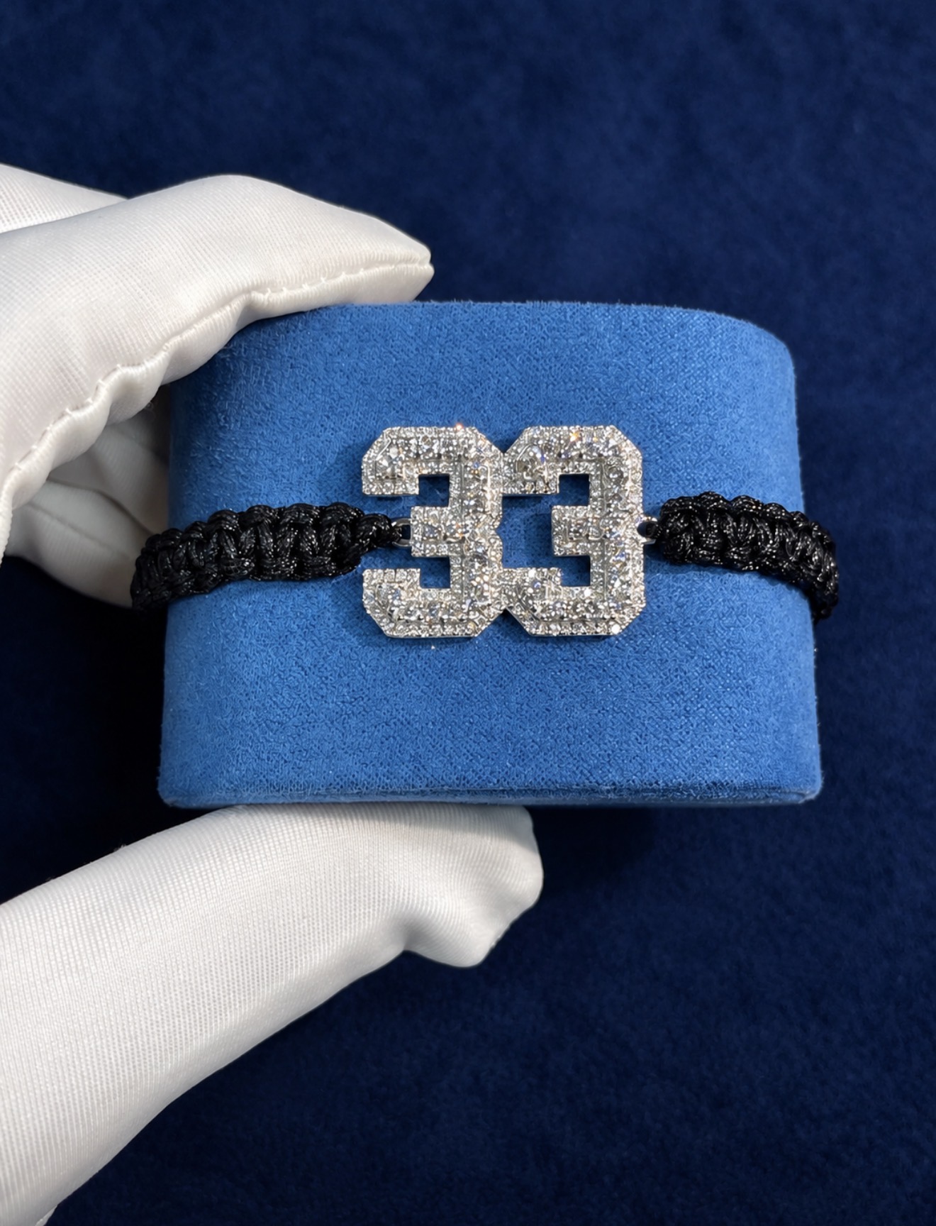 Custom signature bracelet with a paved numeral charm on knotted black cord, held in a white glove against a navy background